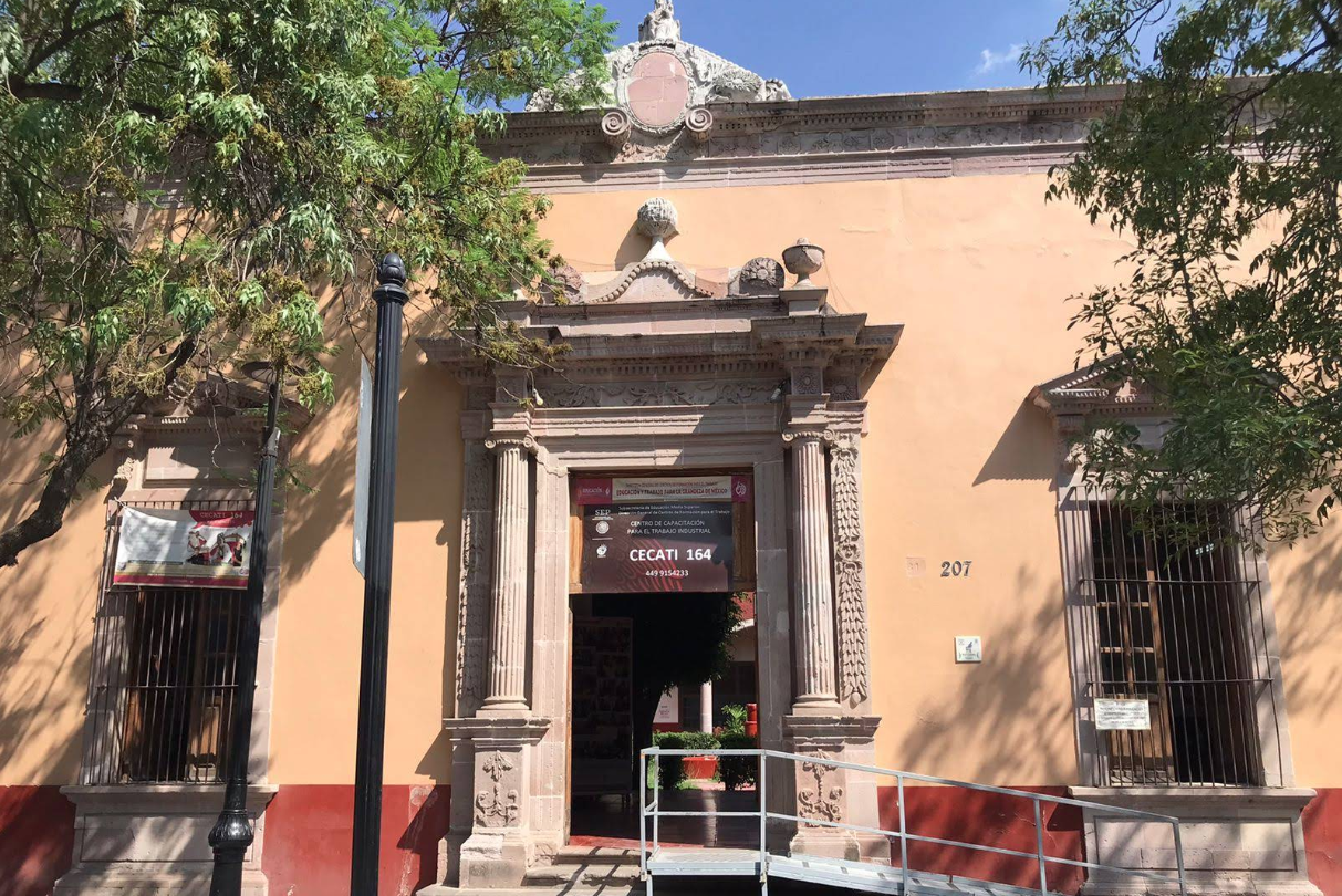 Fotografía donde se muestra la fachada del plantel con paredes color naranja. Al centro hay una puerta con un letrero que dice CECATI 164 y se puede ingresar por medio de escaleras o rampa. 